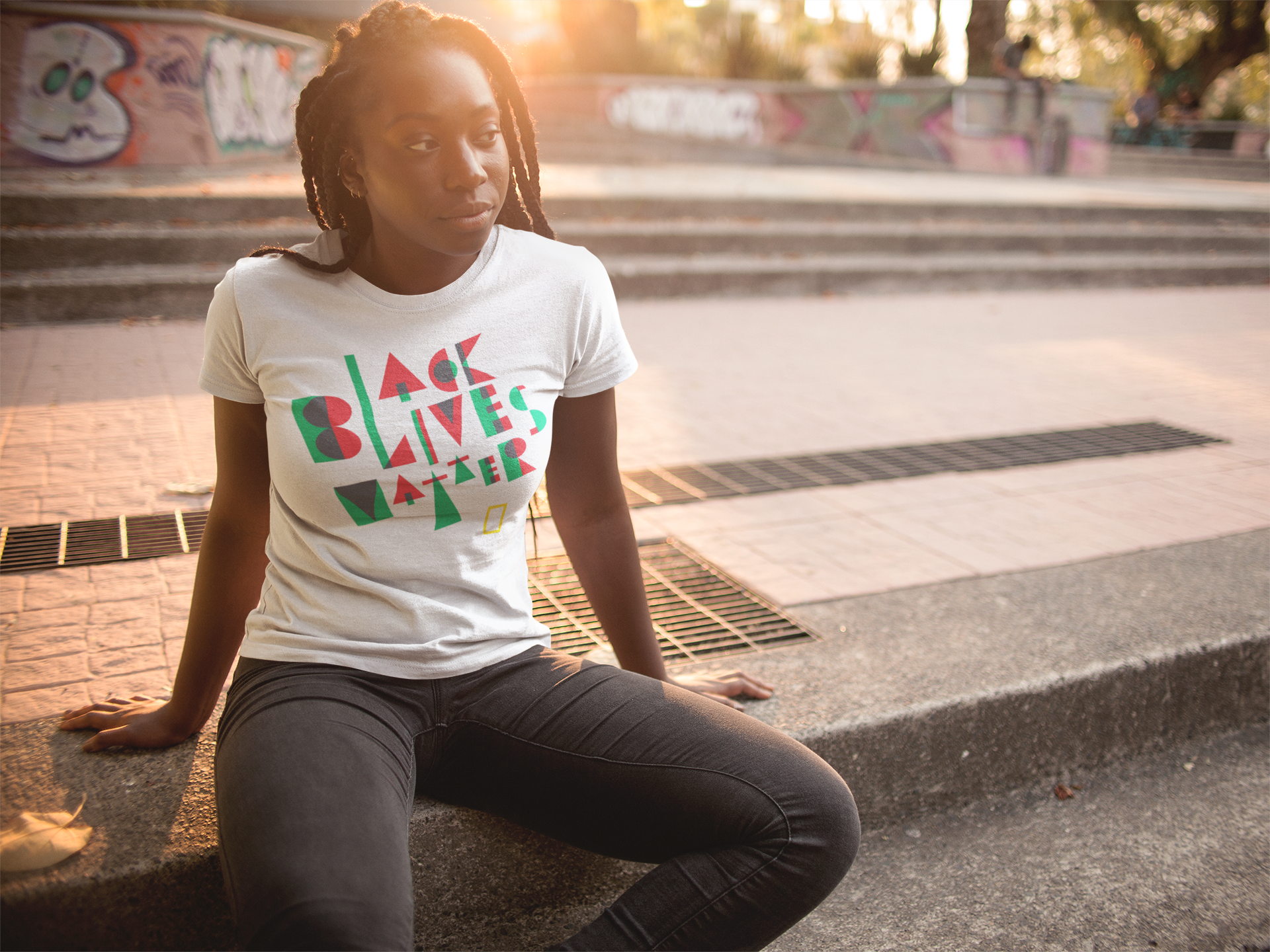 A person with long braided hair sits outdoors on concrete steps, wearing a white T-shirt with Black Lives Matter in colorful geometric letters. The background is an urban setting with graffiti-covered walls and trees.