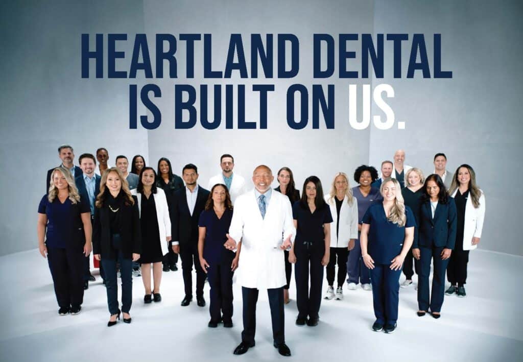 A diverse group of dental professionals stands together, smiling, under text that reads HEARTLAND DENTAL IS BUILT ON US. The central figure is an older person in a white coat, with others in varied professional attire. The background is neutral.